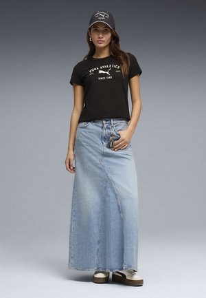 Vrouw met een zwarte Puma Athletics pet en t-shirt, lichtblauwe denim maxi-rok, houdt sleutels vast, staand tegen een grijze achtergrond.