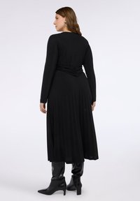 Černé dlouhé šaty s dlouhým rukávem, s přiléhavým živůtkem a nazbieraným zadním dílem, splývavou sukní a hladkou texturou; doplněné černými kotníkovými botami.