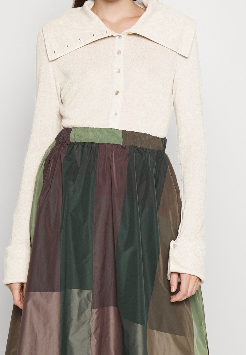 Haut beige à manches longues avec un grand col et une fermeture à boutons, associé à une jupe à couches multicolores dans des tons de vert foncé, marron et olive.