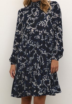 Woman wearing navy blue long-sleeve dress with white floral pattern, high neckline, and tiered skirt, standing against a plain background.