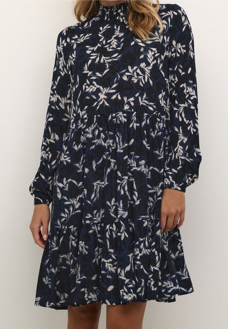 Femme portant une robe bleu marine à manches longues avec un motif floral blanc, un col montant et une jupe à volants, debout devant un fond uni.