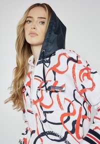 Biała kurtka z czerwonymi i granatowymi abstrakcyjnymi wzorami przypominającymi wir, z granatowym kapturem, zapięciem na zamek i kieszenią z akcentem logo.