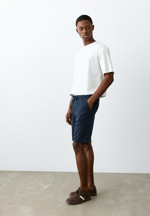 Jeune homme debout, portant un t-shirt blanc à manches courtes, un short cargo bleu marine et des baskets marron, les mains dans les poches, regardant sur le côté.