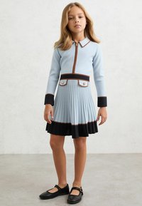 Svetlomodré šaty s dlhými rukávmi, so zipsom vpredu, plisovanou sukňou, kontrastným tmavomodrým lemom, hnedým lemovaním a dvoma ozdobnými gombíkmi na hrudi.