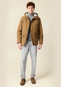 Joven con una chaqueta con capucha color arena sobre un suéter de cuello alto gris, jeans azul claros y zapatos marrones, de pie frente a un fondo beige liso.