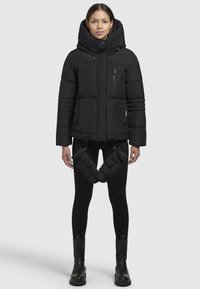Zwarte pufferjack met capuchon, voorzien van een ritszak, gewatteerd design en verstelbare trekkoorden; gecombineerd met zwarte leggings en laarzen.