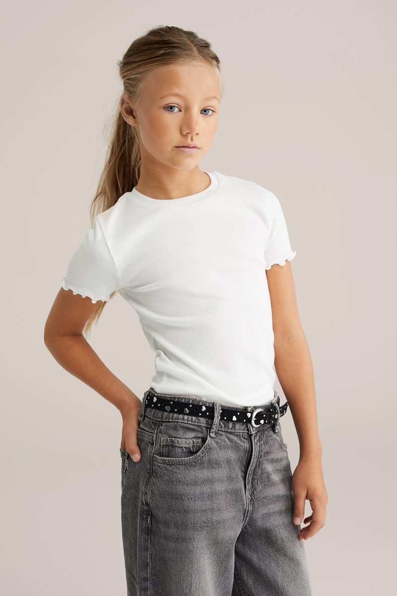 Valge sooniline T-särk, millel on lainelised lühikesed varrukad, koos hallide denimpükstega ja musta vööga, millel on valged täpid.