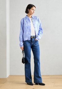 Lyseblå bluse med blomster mønster og knaplukning, parret med mørkeblå flared jeans og sorte høje ankelstøvler, mens hun holder en lille sort taske.