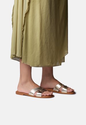 Stopy w metalicznych, złotych płaskich sandałach z podwójnymi paskami, pokazane z lekką, oliwkowozieloną spódnicą sięgającą do połowy łydki.