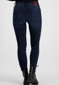 Schmale Jeans aus dunkelblauem Denim. Mit hoher Taille, zwei Gesäßtaschen und einem braunen Lederetikett am Bund. Glatte Textur.
