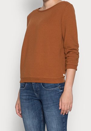 Henkilö, joka pukeutunut rustinsävyiseen, teksturoituun pitkähihainen yläosa ja sinisiin farkkuihin, seisoo yksinkertaisen valkoisen taustan edessä.