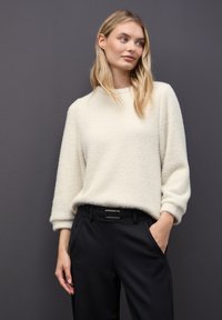 Suéter de punto crema con un acabado texturizado, cuello redondo y corte relajado, combinado con pantalones negros y un cinturón simple.