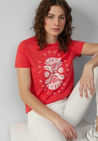 T-shirt en coton rouge avec un graphisme floral blanc et un design textuel, à manches courtes et coupe décontractée. Associé à un pantalon blanc.