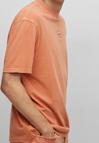 T-shirt a maniche corte, con collo a giro in morbido tessuto arancione chiaro, caratterizzato da una discreta etichetta con logo sul petto e dettagli di cucitura in vista.