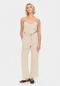 Beige jumpsuit with thin straps, drawstring waist, and wide-leg pants. Smooth fabric, casual design, no patterns or embellishments.