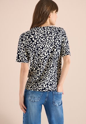 Vrouw met lang bruin haar, gekleed in een zwart-wit shirt met korte mouwen en luipaardprint en een blauwe spijkerbroek, staand met één hand in haar achterzak.