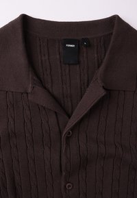 Καφέ πλεκτή ζακέτα με υφή πλεξούδας, διαθέτει γιακά, κλείσιμο με κουμπιά και ετικέτα με την ένδειξη «FORMER» σε μαύρο. Μέγεθος L.