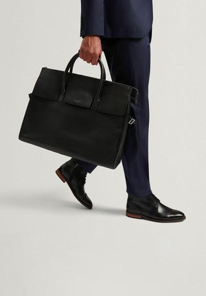 Valigetta in pelle nera dal design strutturato, con manici superiori e texture liscia, portata insieme a scarpe formali scure e pantaloni.