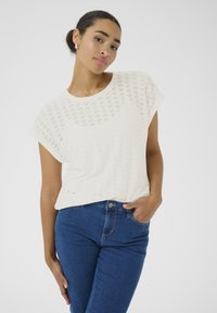 Haut en tricot perforé blanc à manches courtes, col rond et coupe ample, associé à un jean en denim bleu.