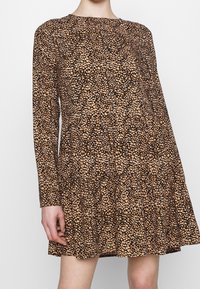 Långärmad klänning ovanför knäet med ett svart och tan oregelbundet prickmönster, figurnära midja och lös kjol, buren av en person som står mot en vit bakgrund.