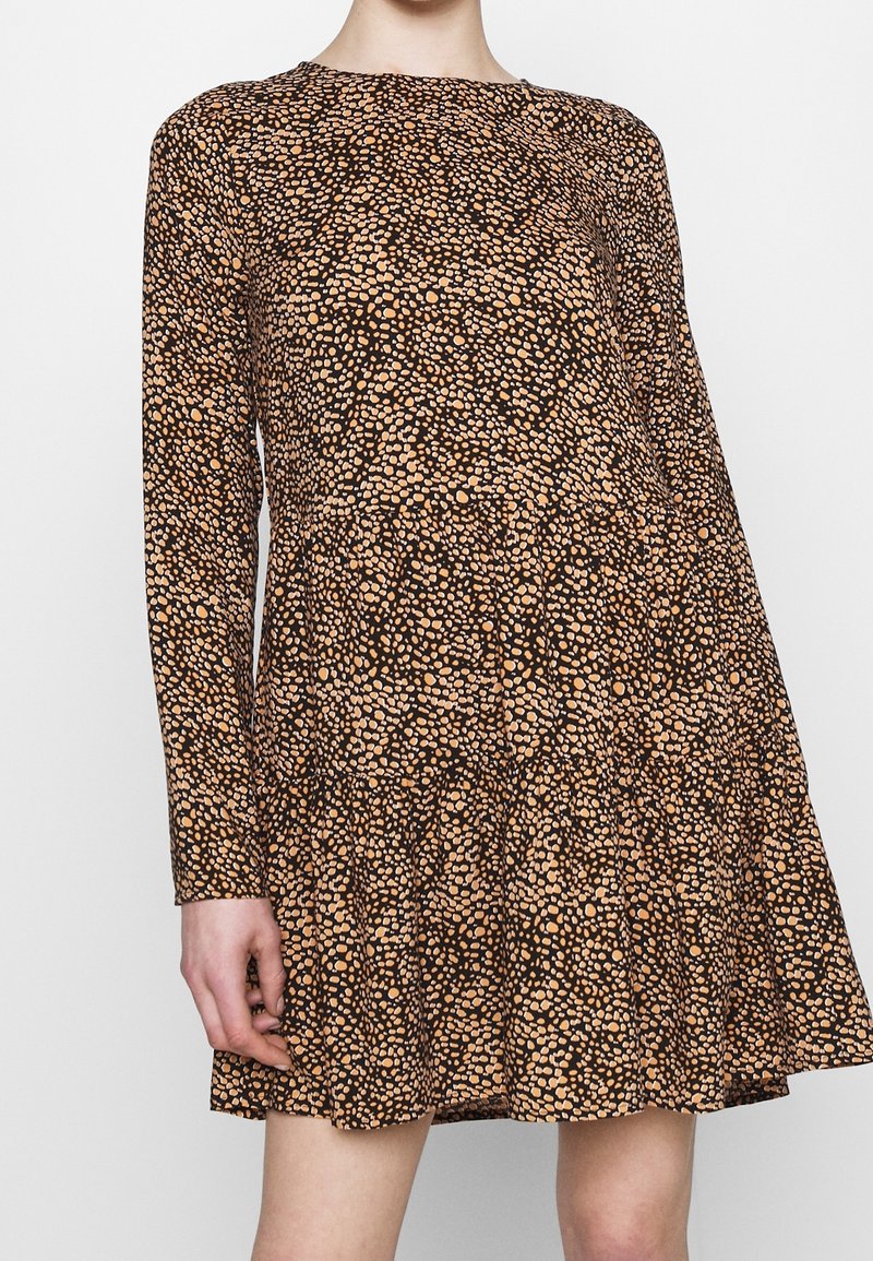 Långärmad klänning ovanför knäet med ett svart och tan oregelbundet prickmönster, figurnära midja och lös kjol, buren av en person som står mot en vit bakgrund.