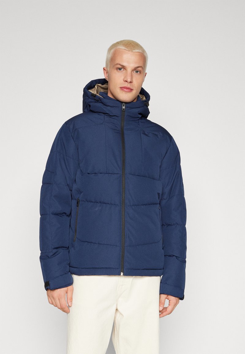 Giacca imbottita blu navale con colletto alto e cappuccio regolabile; presenta chiusura frontale con zip e tasche laterali; texture trapuntata.
