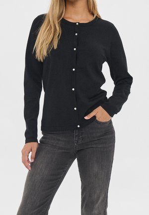 Woman wearing a black long-sleeve cardigan with pearl buttons and dark grey jeans, one hand in pocket, against a plain background.