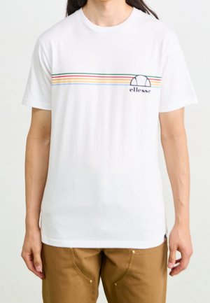 Mężczyzna w białej koszulce z cienkimi poziomymi tęczowymi paskami i małym logo ellesse na piersi, w połączeniu z brązowymi spodniami.