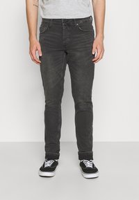 Muž v vybledlých černých slim-fit džínách s ohrnutými nohavicemi, černých plátěných teniskách s bílými tkaničkami a svetru světle šedé barvy.