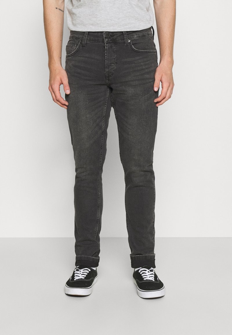 Muž v vybledlých černých slim-fit džínách s ohrnutými nohavicemi, černých plátěných teniskách s bílými tkaničkami a svetru světle šedé barvy.