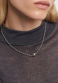 Sølvperlekæde med et centralt rundt charme, der ligger på en mørk, struktureret turtleneck sweater.