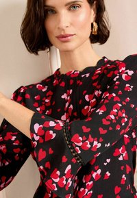 Schwarze Bluse mit roten und rosa Herzmustern, hochgeschnittenem Ausschnitt und Glockenärmel mit Spitzenbesatz. Glatte Stoffstruktur.