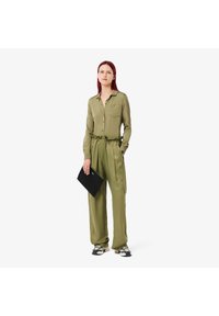 Tuta verde oliva con colletto, vita con cordino e maniche lunghe; il modello tiene una clutch nera e indossa delle sneakers.