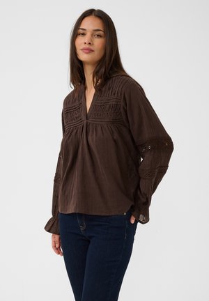 Frau mit langen dunklen Haaren, die eine lockere dunkelbraune Bluse mit strukturierten Details und dunkelblaue Jeans vor einem schlichten hellen Hintergrund trägt.