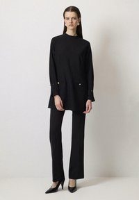 Blusa negra de manga larga con detalles de botones dorados y pantalones rectos, combinados con tacones negros de punta; tejido suave y corte relajado.