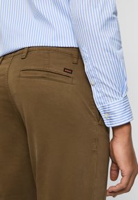 Pantalones de algodón marrón con corte ajustado, que cuentan con un bolsillo trasero y una etiqueta de logo. Combinados con una camisa de botones a rayas azul claro.