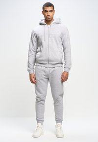 Sudadera con capucha gris y cremallera, pantalones de chándal a juego y zapatillas blancas de caña alta. Presenta puños acanalados y un logo en el pecho.