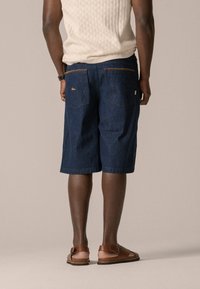 Shorts en denim bleu foncé, de longueur mi-cuisse, avec des poches arrière à couture contrastée et un petit logo. Des sandales marron complètent la tenue.