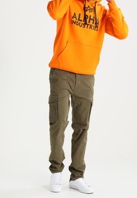 Felpa arancione con cappuccio e logo nero "ALPHA INDUSTRIES", abbinata a pantaloni cargo oliva e sneakers bianche, che mostrano tasche e un taglio casual.