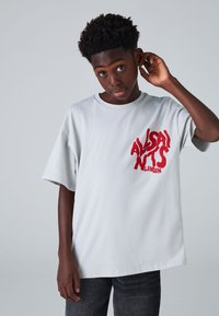 Lysegrå kortærmet T-shirt med et rødt grafisk tekstlogo, der lyder "ALLSAINTS LONDON." Blødt, glat materiale, afslappet pasform.