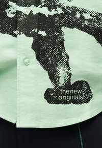 Chemise vert clair avec un graphisme pixellisé noir et le texte « the new originals » près du bas, montrant des boutons et une partie d'un pantalon foncé en dessous.
