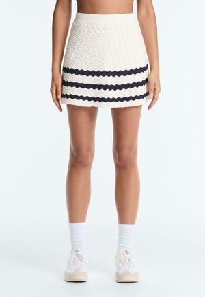 Žena v bielej textúrovanej mini sukni s tromi horizontálnymi čiernymi vlnkovanými pruhmi, bielych ponožkách a bielych teniskách, stojaca na bielom pozadí.