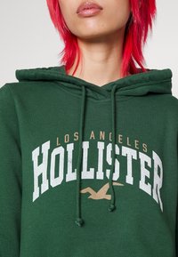 Sudadera con capucha verde con texto blanco y dorado "HOLLISTER" y "LOS ÁNGELES". Presenta cordones y una textura suave.