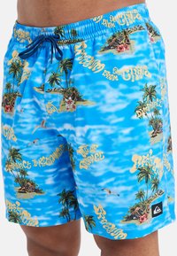 Modré plavky s tropickými grafikami ostrova, palmami a žltým textom. Dizajn má ľahkú a hladkú textúru.