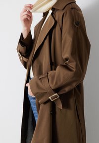 Bruine trenchcoat met een ceintuur in de taille, grote revers, knoopaccenten en een relaxte pasvorm. Draag het over een witte trui en blauwe jeans.