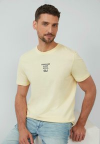 Gelbes, kurzärmeliges T-Shirt mit Rundhalsausschnitt. Schwarzer Text "LONESOME RIDER SOCIAL Club" auf der Brust aufgedruckt. Weiches Baumwollgewebe.