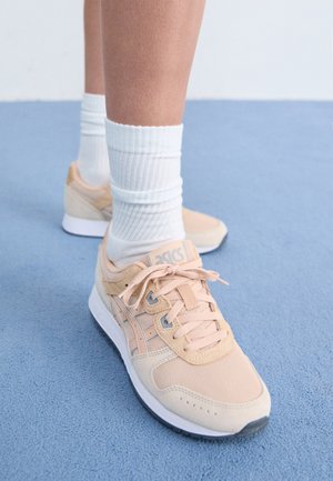 Jasnopomarańczowe buty sportowe z tekstylnym wierzchem, białą gumową podeszwą i sznurowanym zapięciem. Połączone z białymi skarpetami ściąganymi i ustawione na niebieskiej powierzchni.