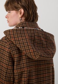 Bruine geruite jas met een capuchon, met een ruitpatroon in tinten bruin, zwart en subtiele rode accenten. Gestructureerde stofafwerking.