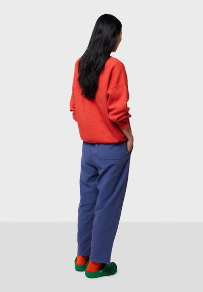 Rød oversized sweatshirt, blå løstsiddende bukser, grønne træsko og orange sokker. Bløde teksturer og afslappet design, set fra bagsiden.
