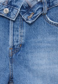 Vaaleansiniset farkut, joissa on nappikiinnitys, oranssia ommelta ja haalistunut pinta. Kankaan pinnassa on pieniä kulumia.